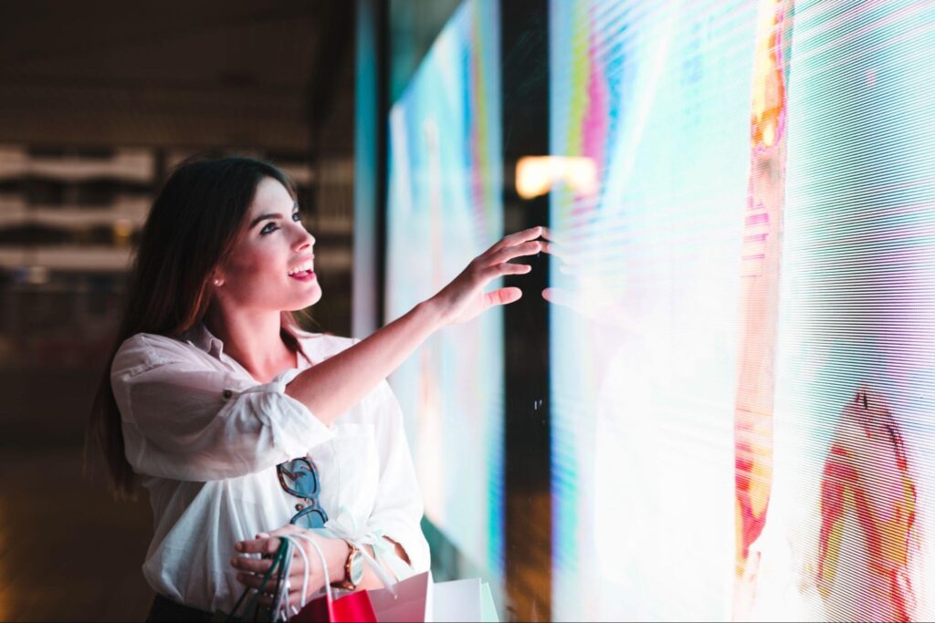 A smiling woman with shopping bags reaches out to touch a large, brightly lit digital display screen