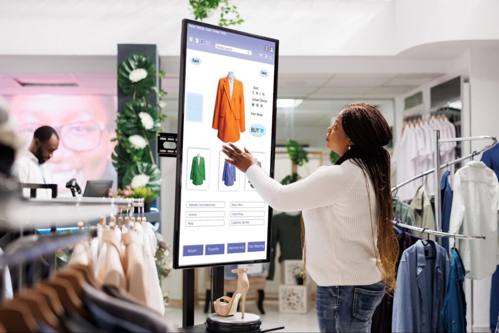 A woman in a clothing store touches an interactive digital screen displaying a jacket for sale, with options to choose sizes, colors, and buy online