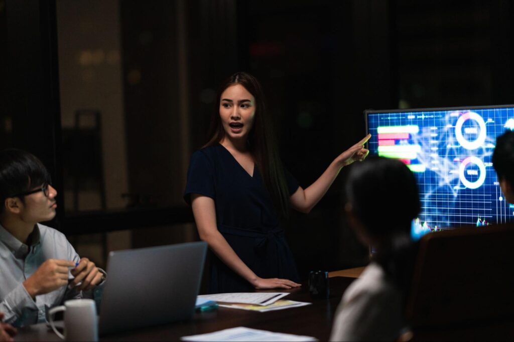 A woman gestures toward a large digital screen displaying charts and data during a business meeting in a dim office